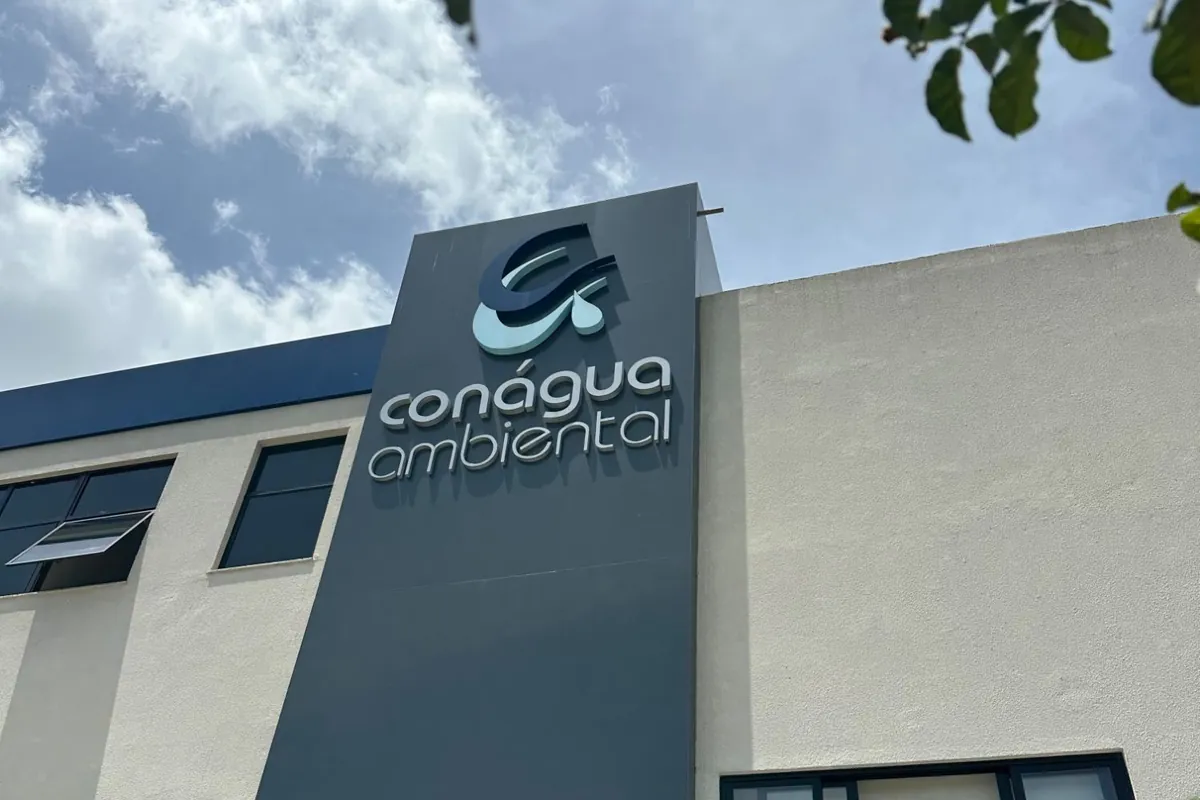 Fachada da sede da Conágua Ambiental, com o letreiro da empresa, vista de baixo entre galhos de árvore
